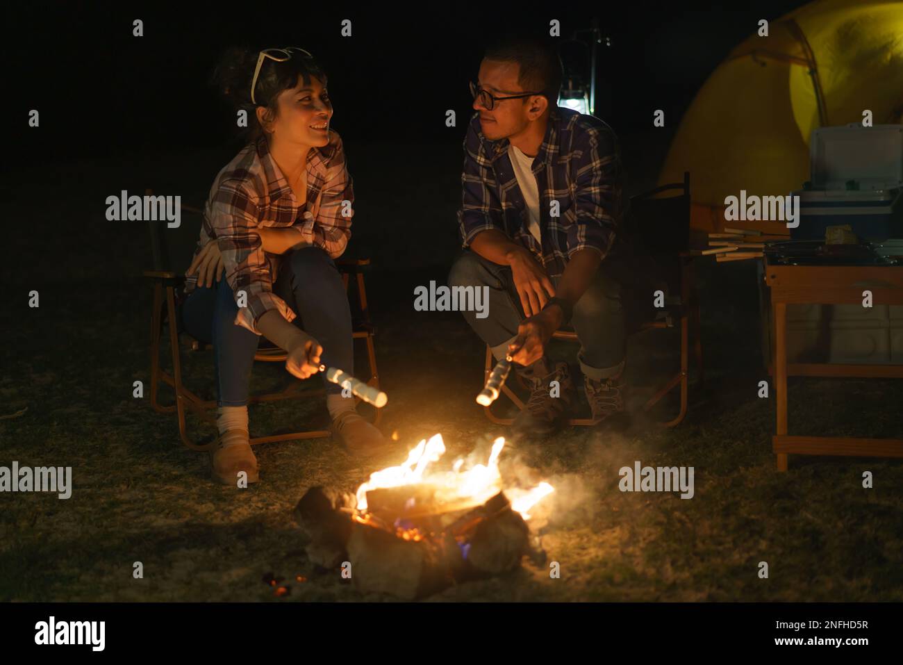 Un couple asiatique torréfaction de guimauves ensemble à un feu de camp où ils ont installé une tente pour camper au bord du lac la nuit. Banque D'Images