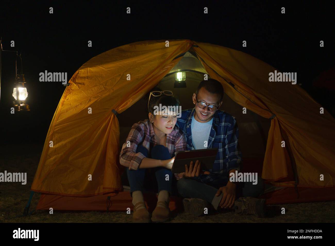 Le couple asiatique joue sur sa tablette numérique dans sa tente pendant la nuit en planifiant un voyage pour demain. Banque D'Images