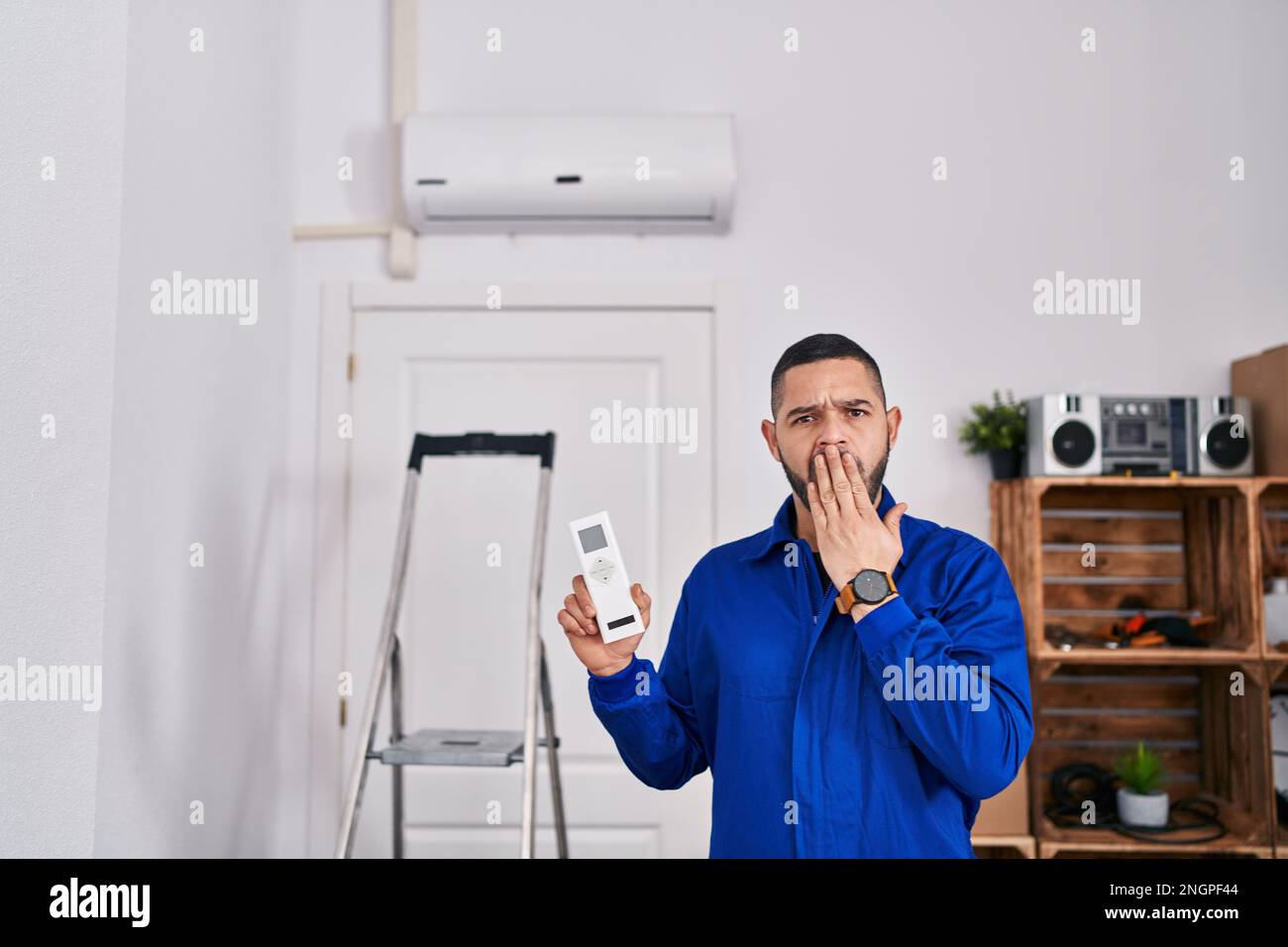 Réparateur hispanique travaillant avec un climatiseur couvrant la bouche avec la main, choqué et peur de l'erreur. expression surprise Banque D'Images