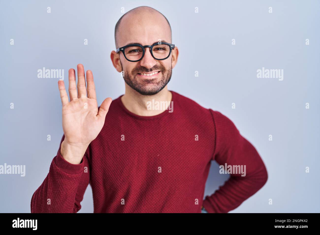 Jeune homme chauve avec une barbe debout sur fond blanc, portant des lunettes pour dire bonjour heureux et souriant, geste de bienvenue amical Banque D'Images