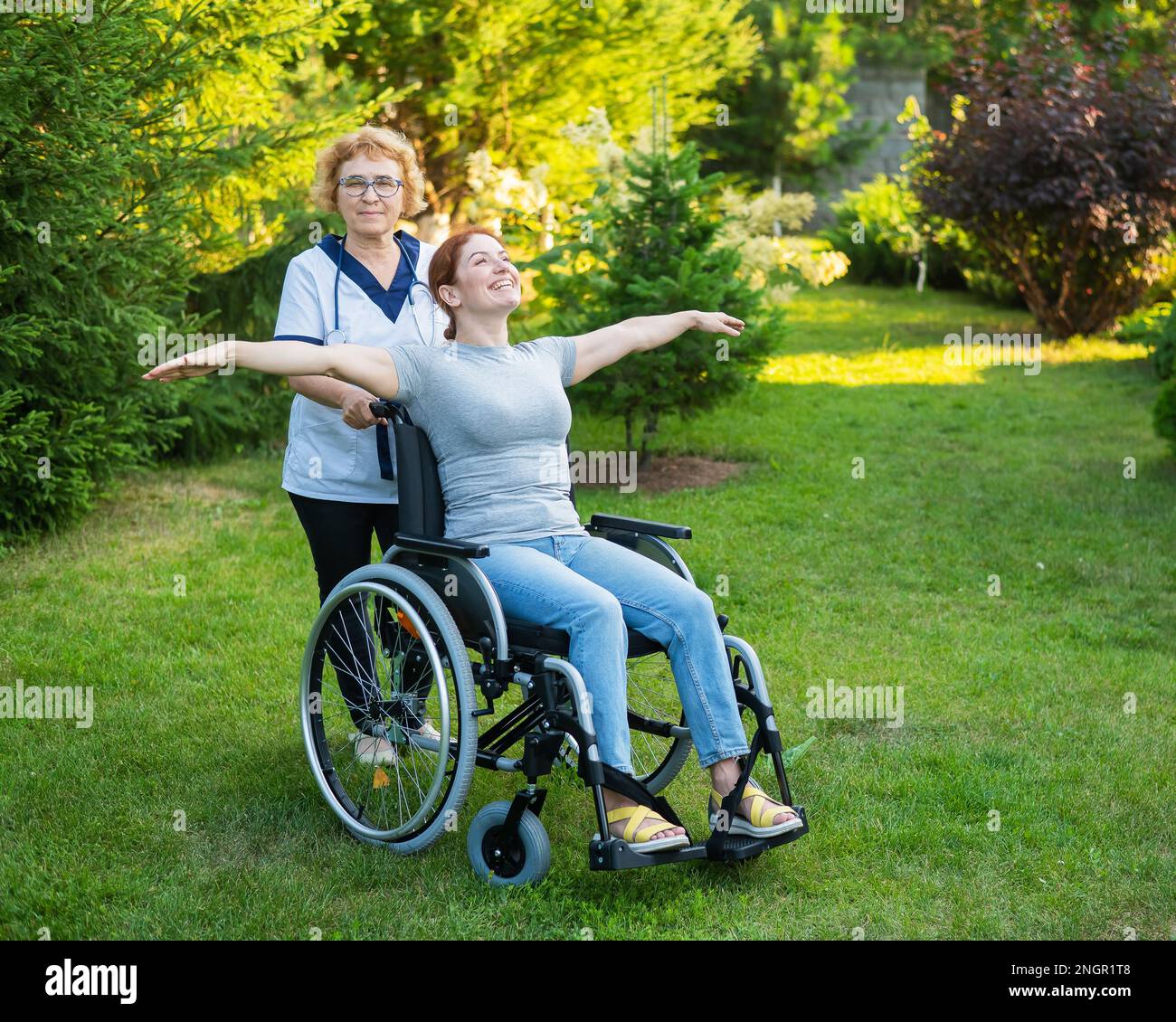 Une infirmière âgée marche avec une femme d'âge moyen en fauteuil roulant dans le parc. La jeune fille a étendu ses bras comme des ailes. Banque D'Images