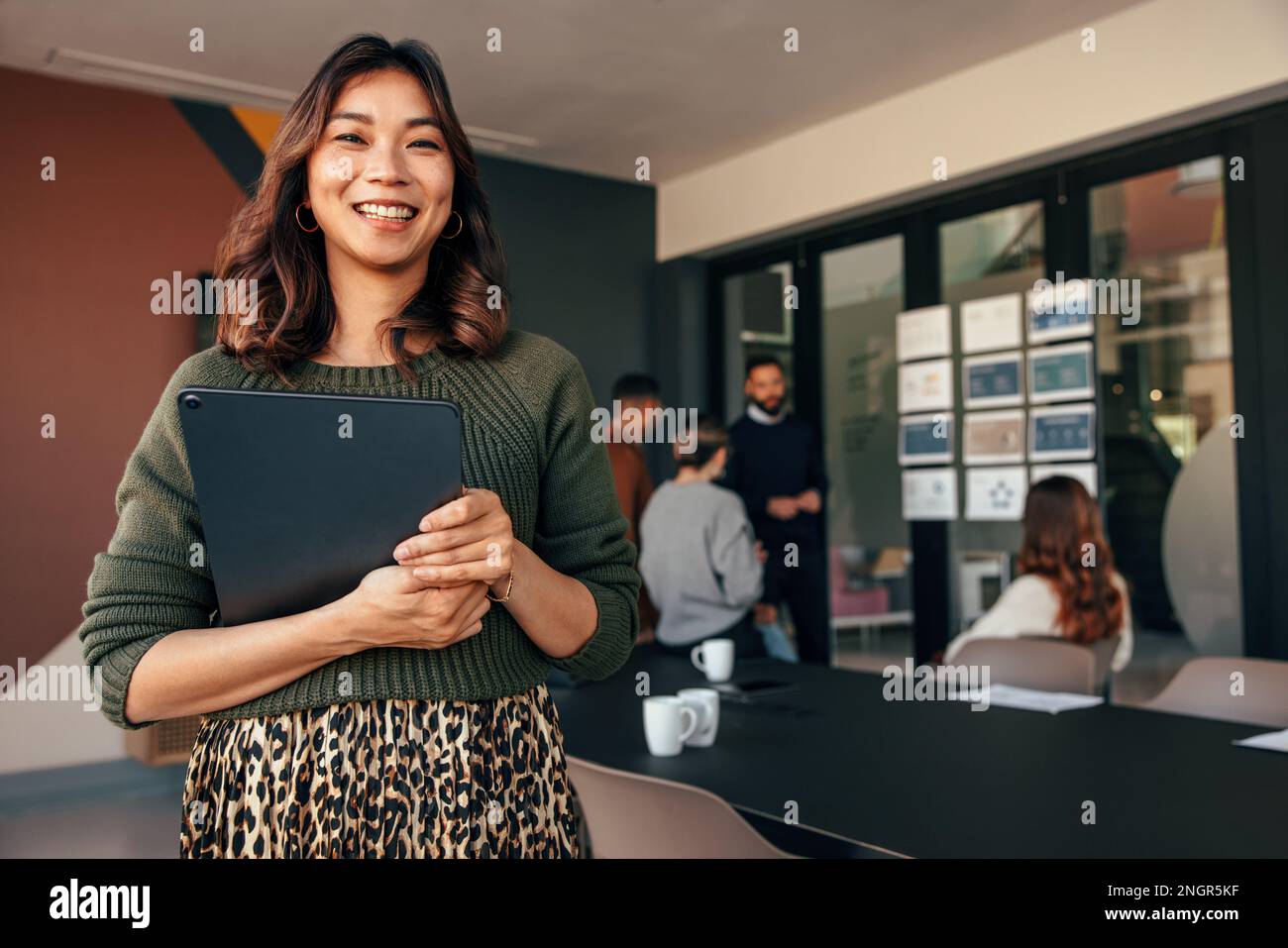 Une jeune femme d'affaires souriante à l'appareil photo tout en tenant une tablette numérique. Bonne jeune femme d'affaires debout dans une salle de réunion avec son collègue Banque D'Images