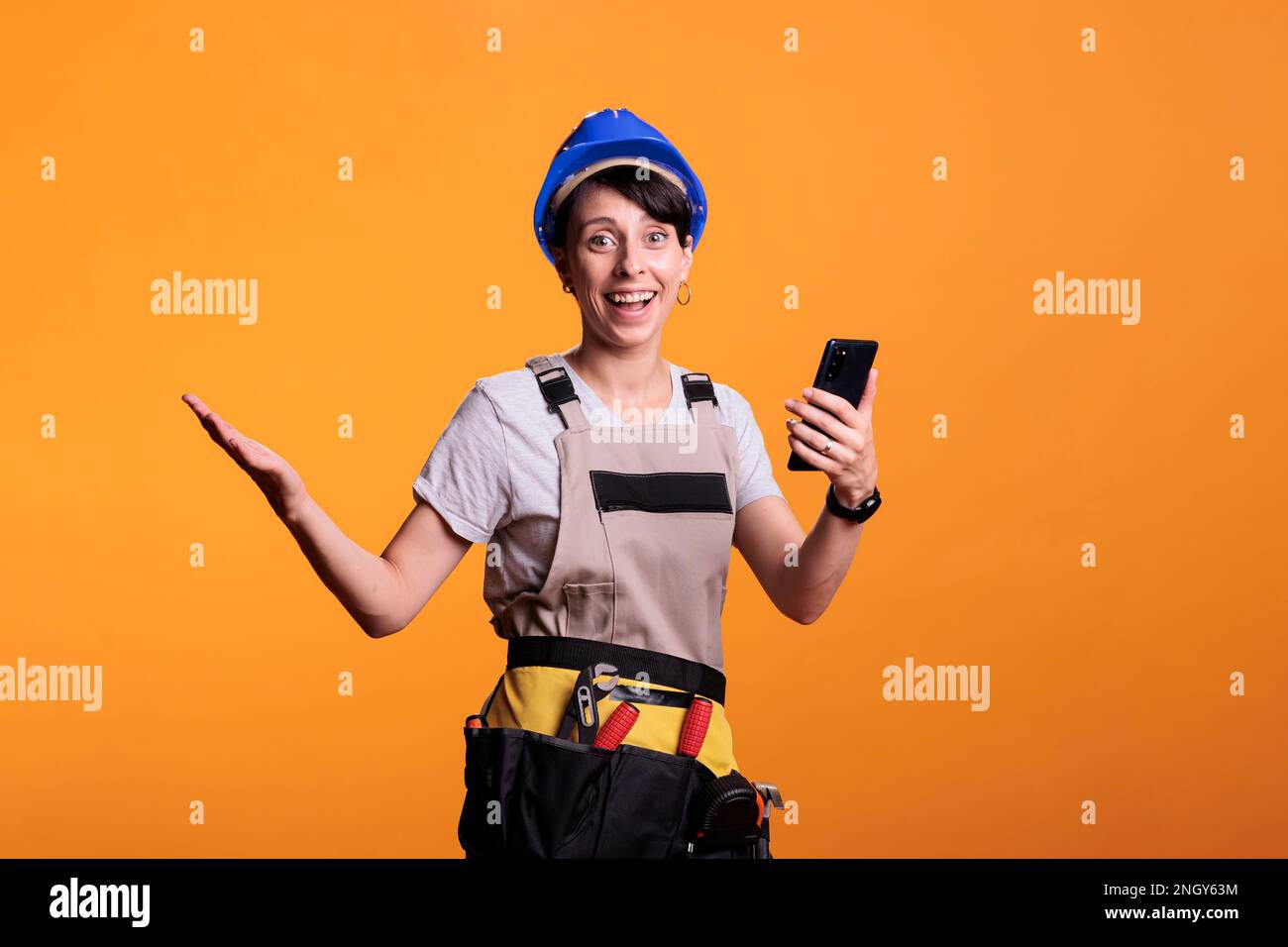 Un ouvrier du bâtiment souriant à l'application pour smartphone, surfant sur Internet et posant en studio. Être heureux et regarder le site Web de téléphone mobile, porter un casque et rénover des combinaisons. Banque D'Images