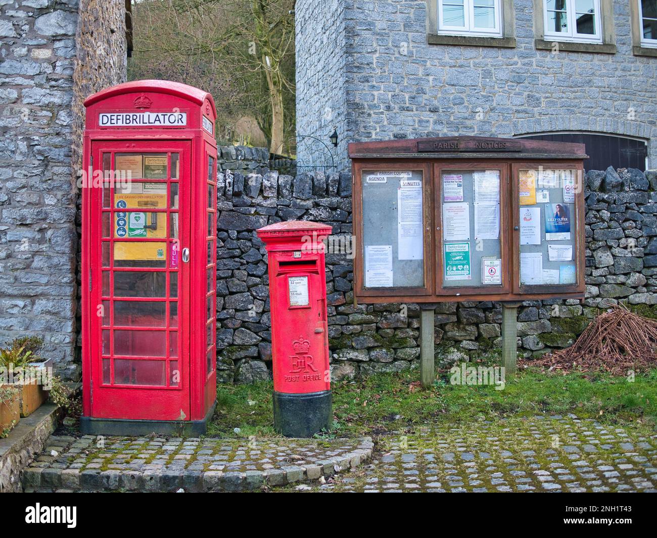 Un défibrillateur d'urgence dans une ancienne boîte téléphonique rouge dans le village de Taddington, dans le Derbyshire, au Royaume-Uni. Une boîte postale rouge et un panneau d'avis paroissial apparaissent o Banque D'Images