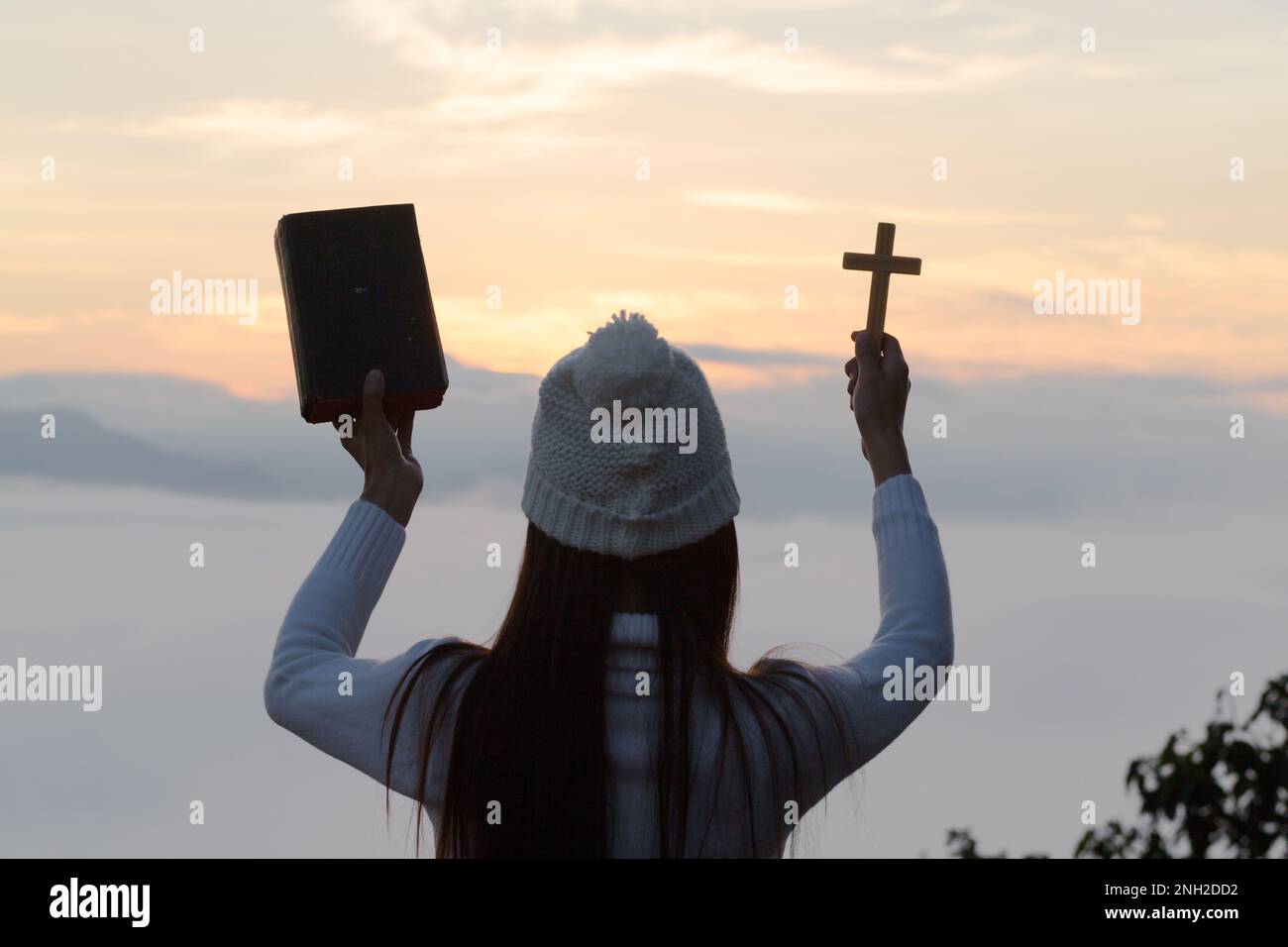Les mains de la jeune femme tiennent la sainte Bible et la levée de la croix chrétienne avec un arrière-plan de coucher de soleil clair. Banque D'Images