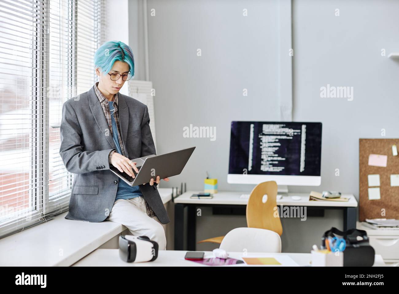 Portrait d'un jeune homme avec des cheveux bleus colorés à l'aide d'un ordinateur portable tout en étant assis près de la fenêtre dans le bureau, espace de copie Banque D'Images