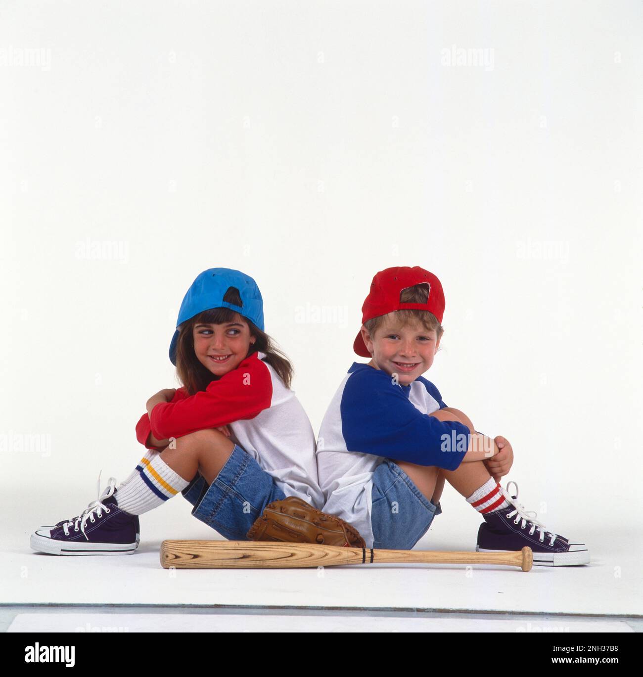 Petit garçon et fille en maillots de baseball les baskets et les casquettes de baseball tournent vers l'arrière assis dos à dos sur un fond blanc avec des gants et une batte de baseball en premier plan Banque D'Images