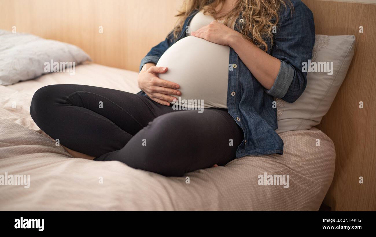 Femme enceinte assise sur un lit tenant son ventre Banque D'Images