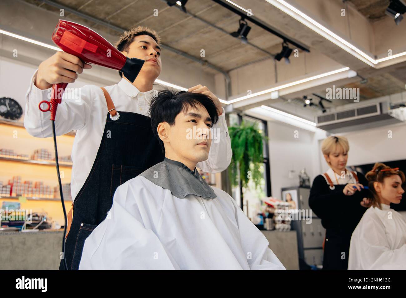 Coiffeur asiatique jeune homme portant un tablier séchant les cheveux de jeune homme beau dans un salon de coiffure moderne tout en se concentrant sur le travail Banque D'Images