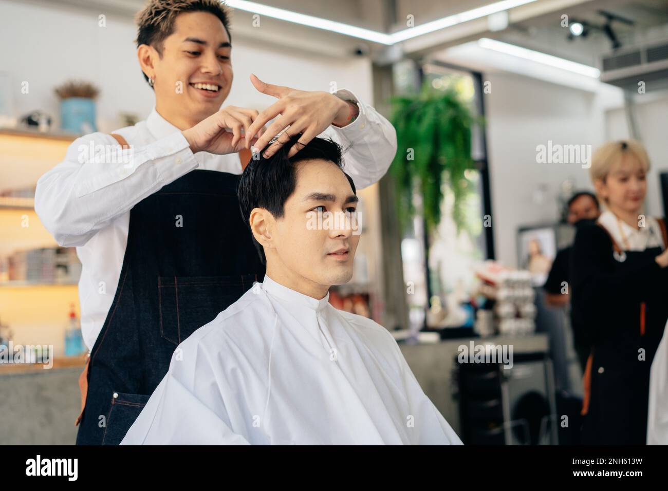 Joyeux coiffeur de jeune asiatique souriant portant un tablier dans moderne salon tenant la brosse et les ciseaux de couper les cheveux de client Banque D'Images