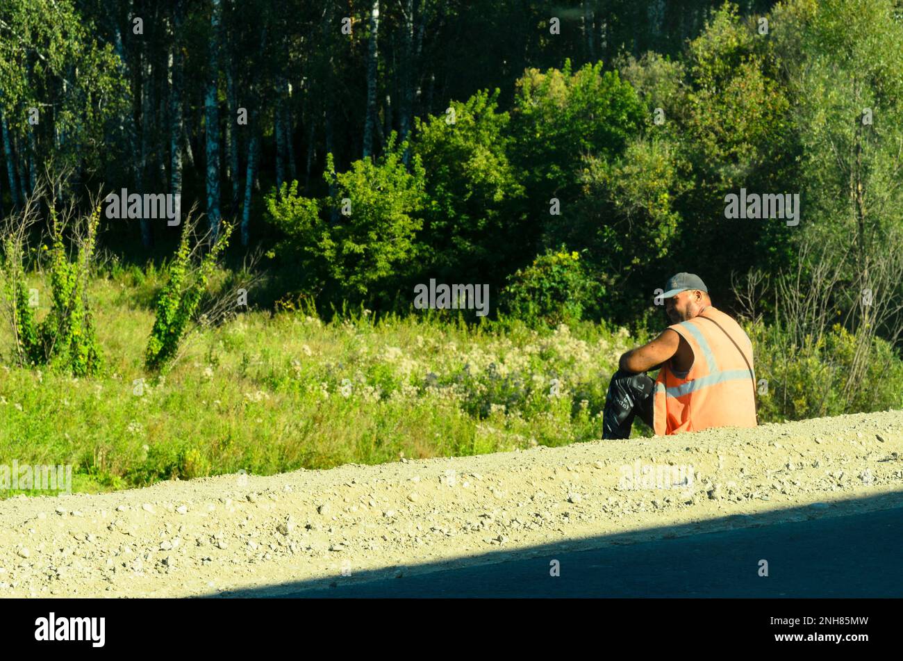 Triste immigrant ouvrier de construction de route dans un gilet orange et chapeau assis sur le côté de la route de la forêt verte et regardant dans la direction Banque D'Images