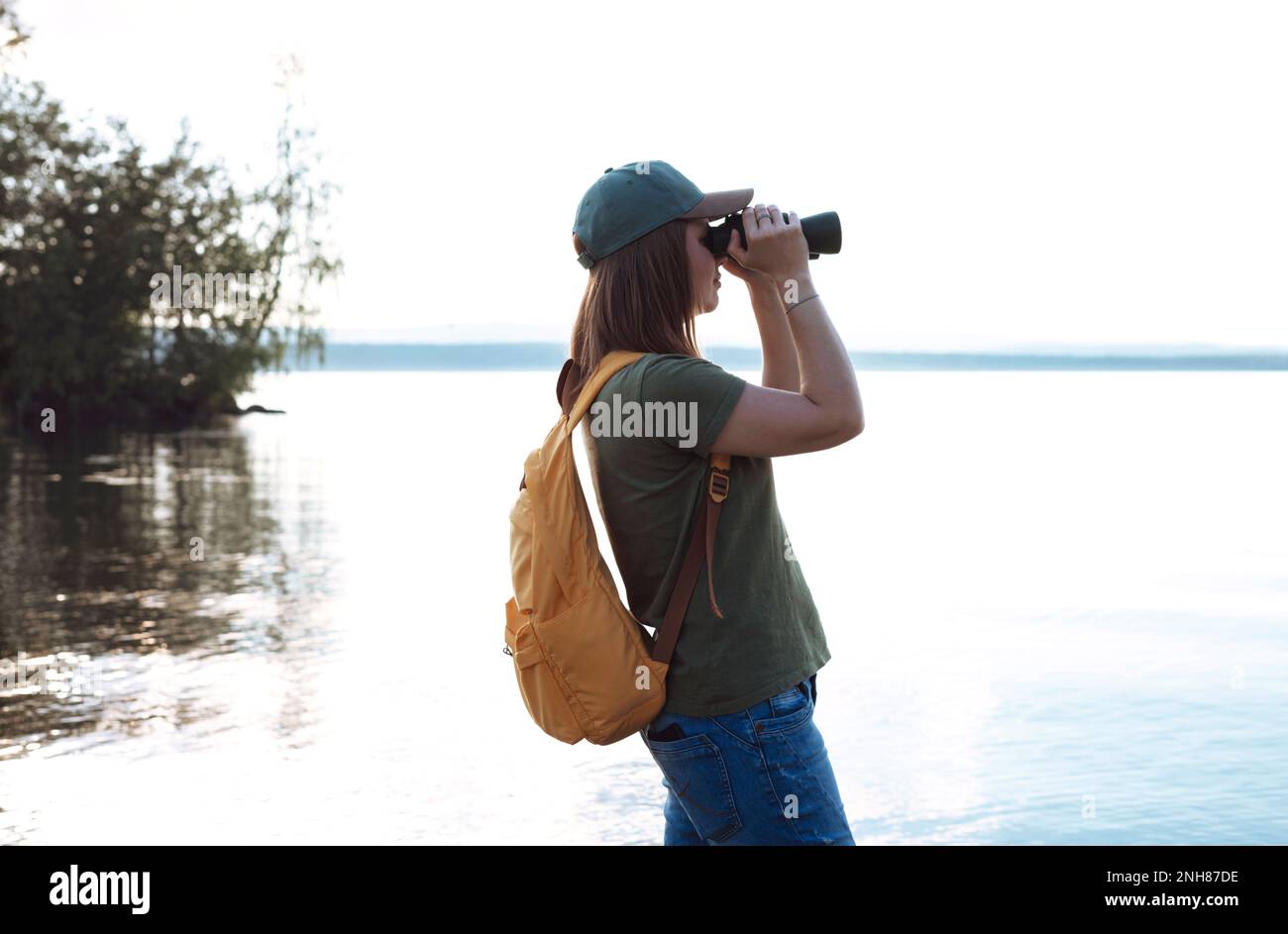 Jeune femme avec sac à dos jaune regardant à travers des jumelles aux oiseaux sur le lac ou la rivière observation des oiseaux, zoologie, écologie recherche dans la nature, observation o Banque D'Images