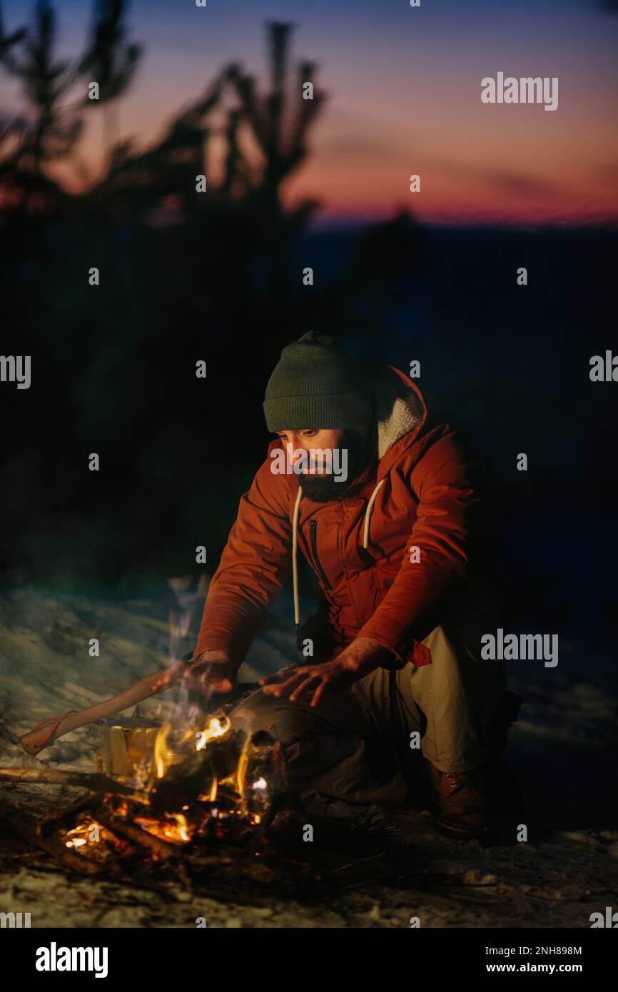 Un homme est assis près d'un feu de camp, se réchauffant par le feu dans une forêt d'hiver au coucher du soleil. Banque D'Images