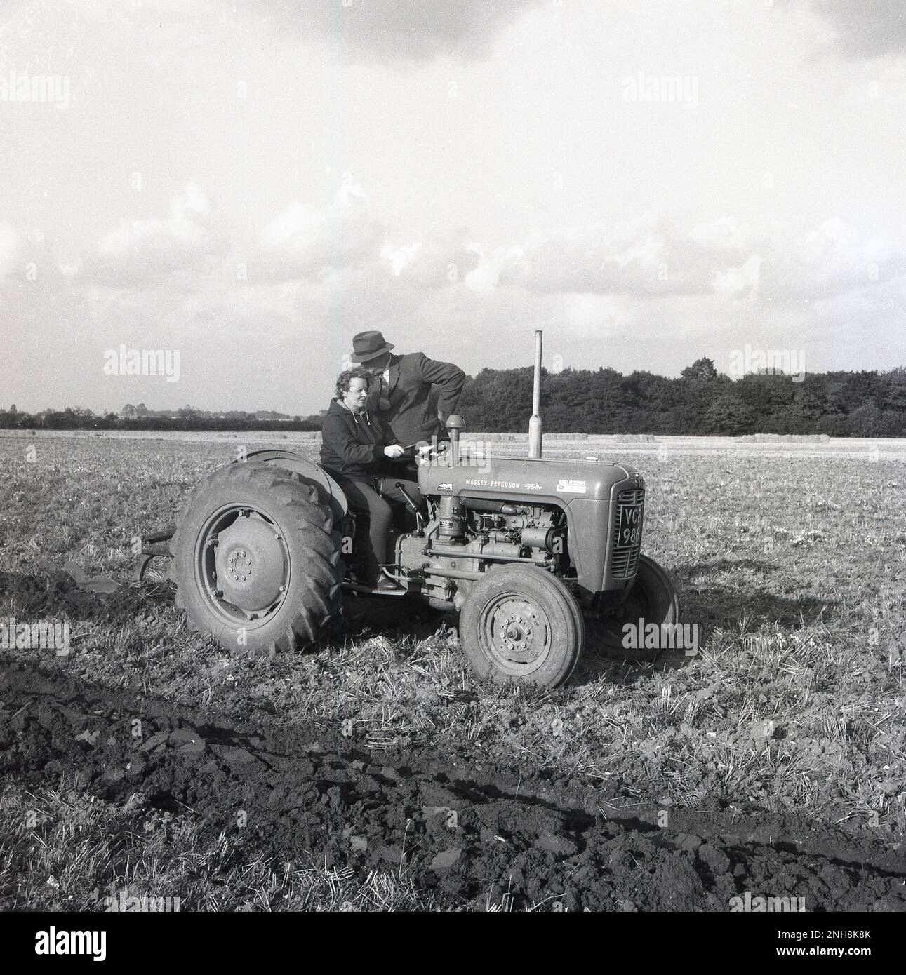 1965, historique, fermier, dans un t-shirt et une cravate, une veste et un chapeau debout sur un tracteur à côté d'une femme pilote, Barton, Angleterre, Royaume-Uni. La femme a une leçon sur la façon de labourer un champ. Le tracteur est un Massey-Ferguson 35X, un modèle classique de l'époque. Banque D'Images