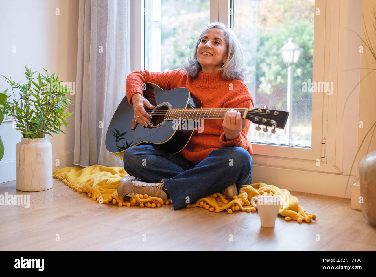 Femme senior avec cheveux blancs jouant la guitare assis sur le sol à la maison et appréciant le chant. Concept: Musique, guitare, chant Banque D'Images