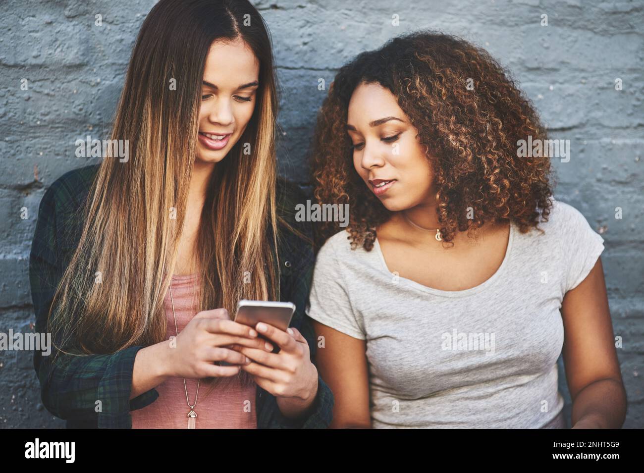 Comment les jeunes d'aujourd'hui suivent les tendances actuelles. deux jeunes femmes debout à l'extérieur et utilisant un téléphone portable contre un mur gris. Banque D'Images