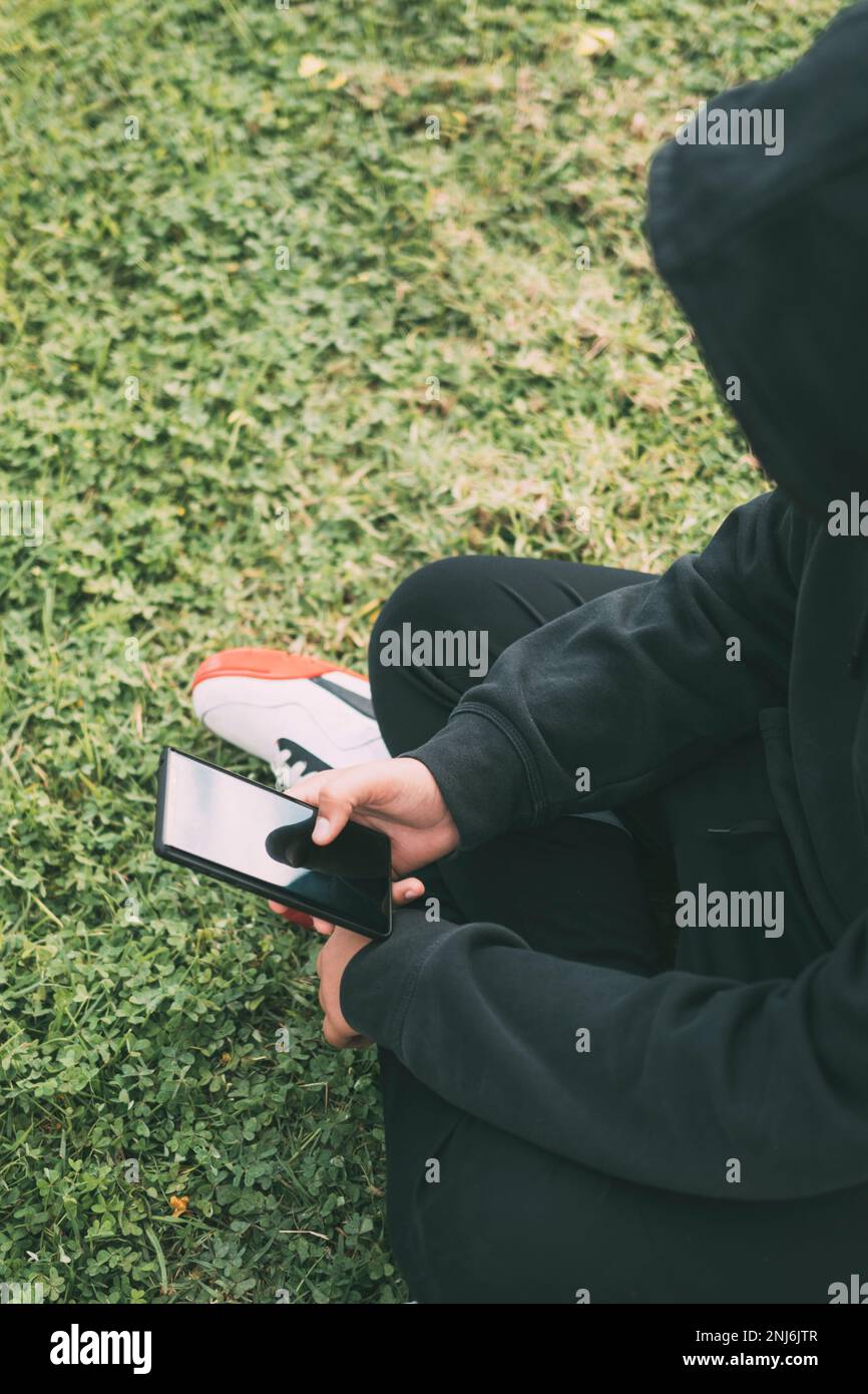 jeune homme assis au sol dans le parc, complètement immergé dans son smartphone. Banque D'Images