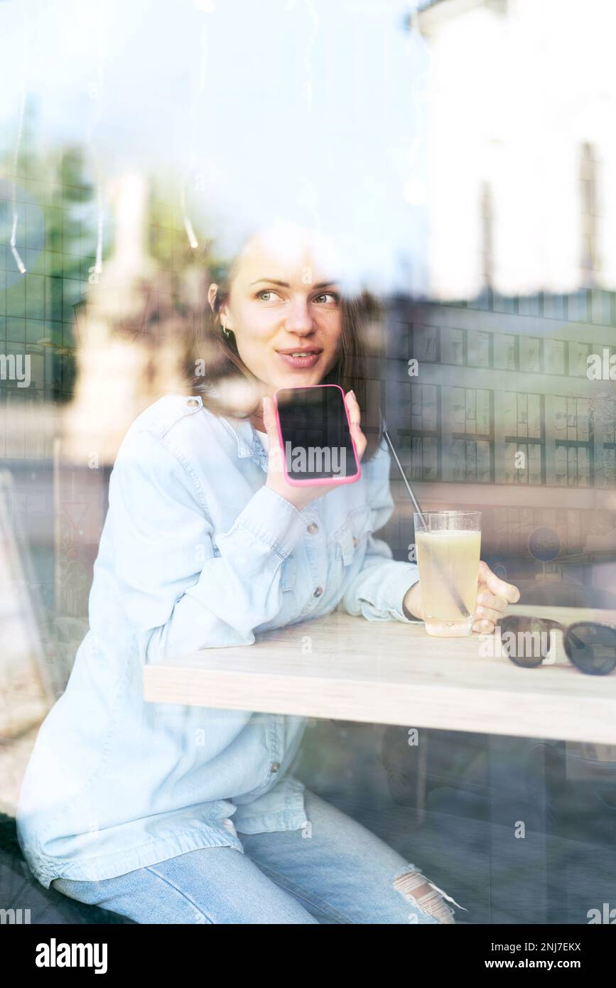 Jeune femme urbaine assise dans un café et enregistrant un message vocal à l'aide de son téléphone portable. Banque D'Images