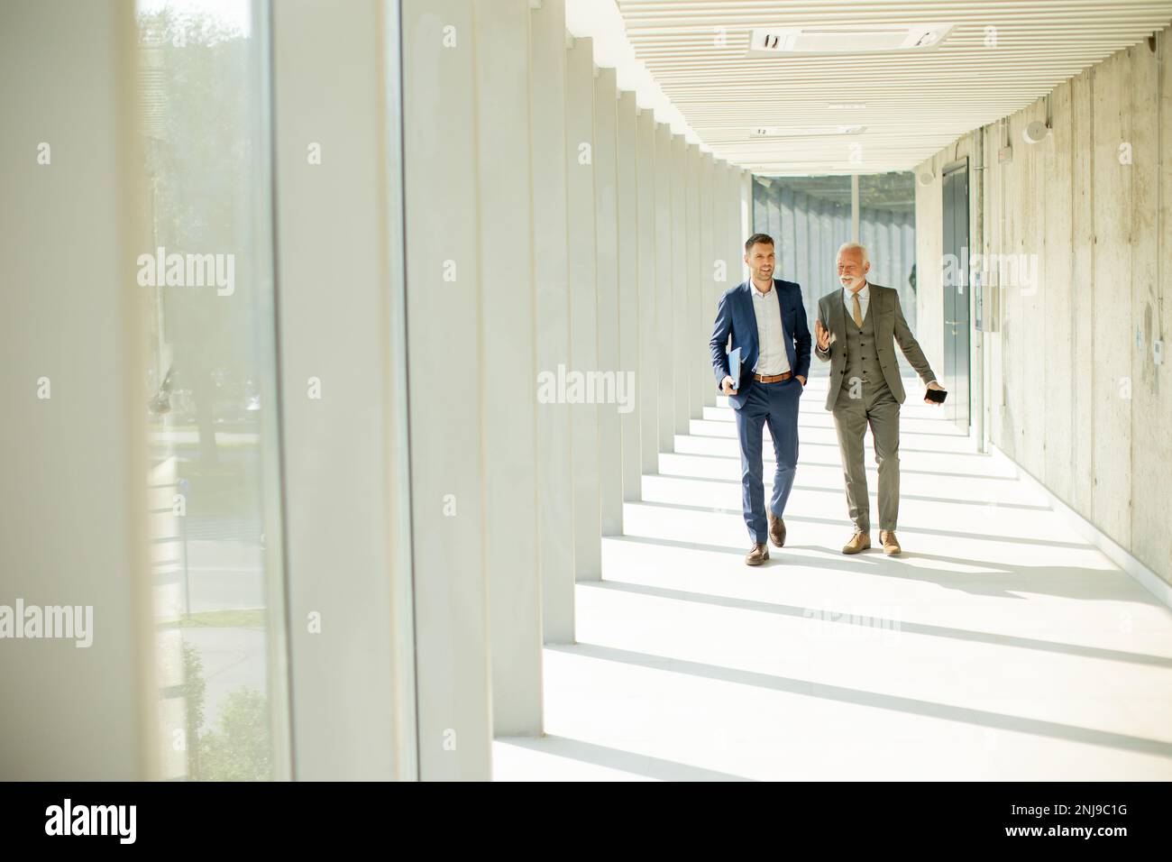 Un jeune homme d'affaires et un homme d'affaires senior descendent dans le couloir d'un bureau, profondément dans la conversation. Ils sont tous deux habillés professionnellement, reflétant leur acum d'affaires Banque D'Images