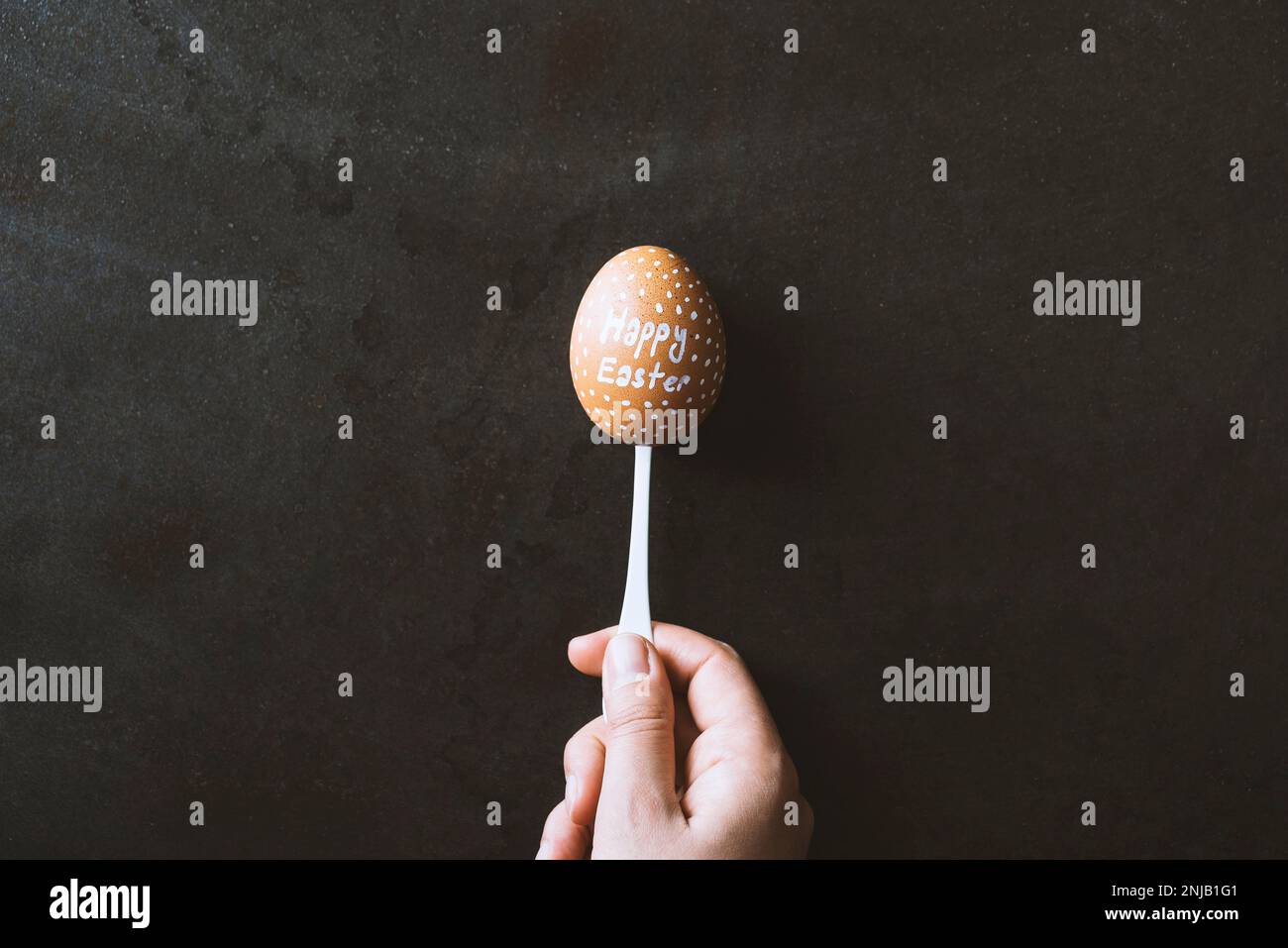 Main tenant un œuf écrit « joyeuses Pâques » dans une cuillère sur fond noir en métal. Banque D'Images