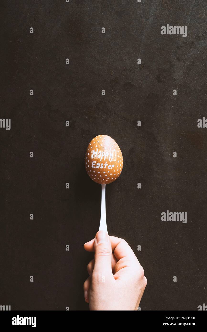 Main tenant un œuf écrit « joyeuses Pâques » dans une cuillère sur fond noir en métal. Banque D'Images