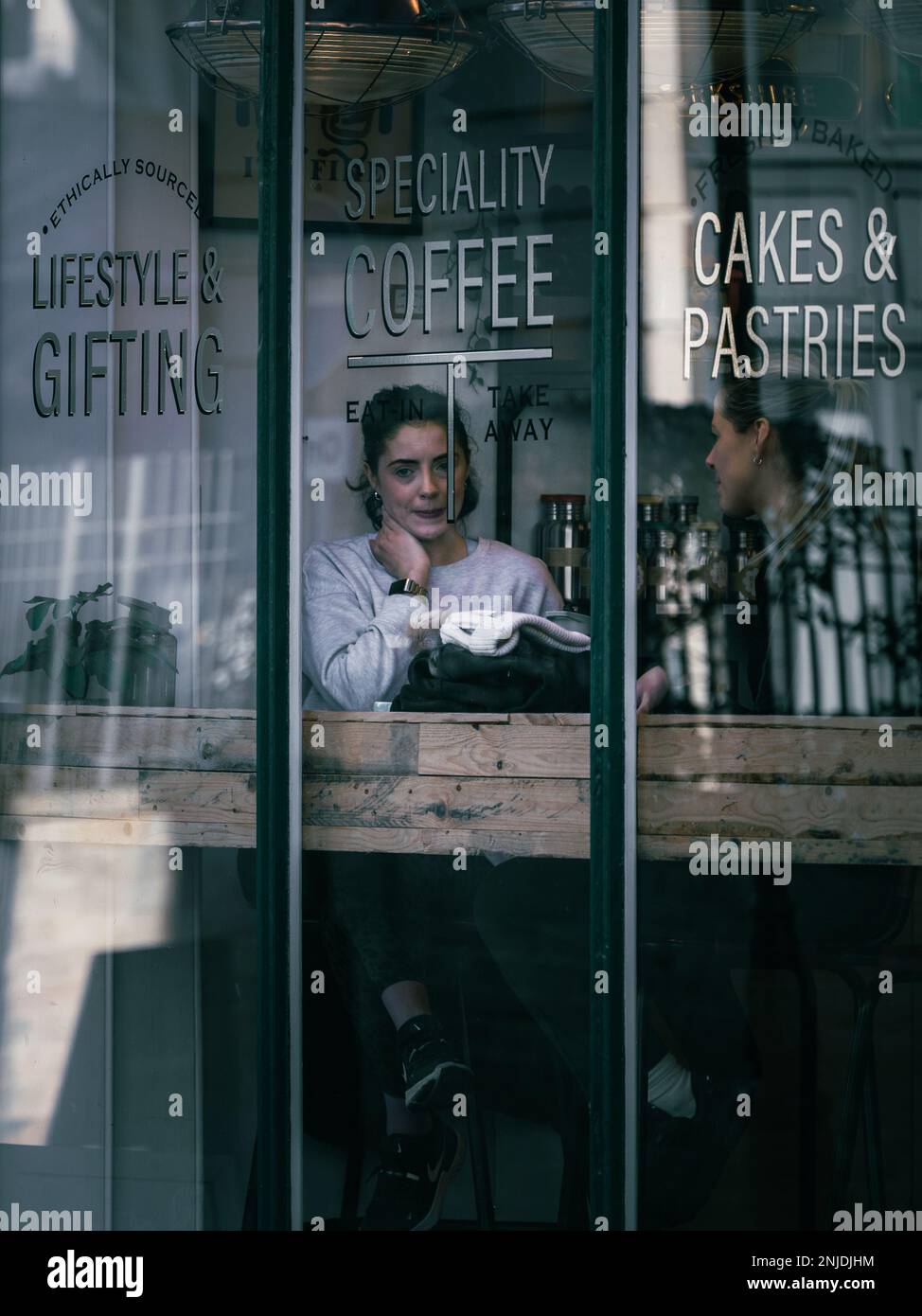 Deux femmes partagent un moment de café tranquille par la fenêtre d'un café confortable, entouré de textures de bois chaudes et de signalétique lettrée à la main offrant une spécialité cof Banque D'Images