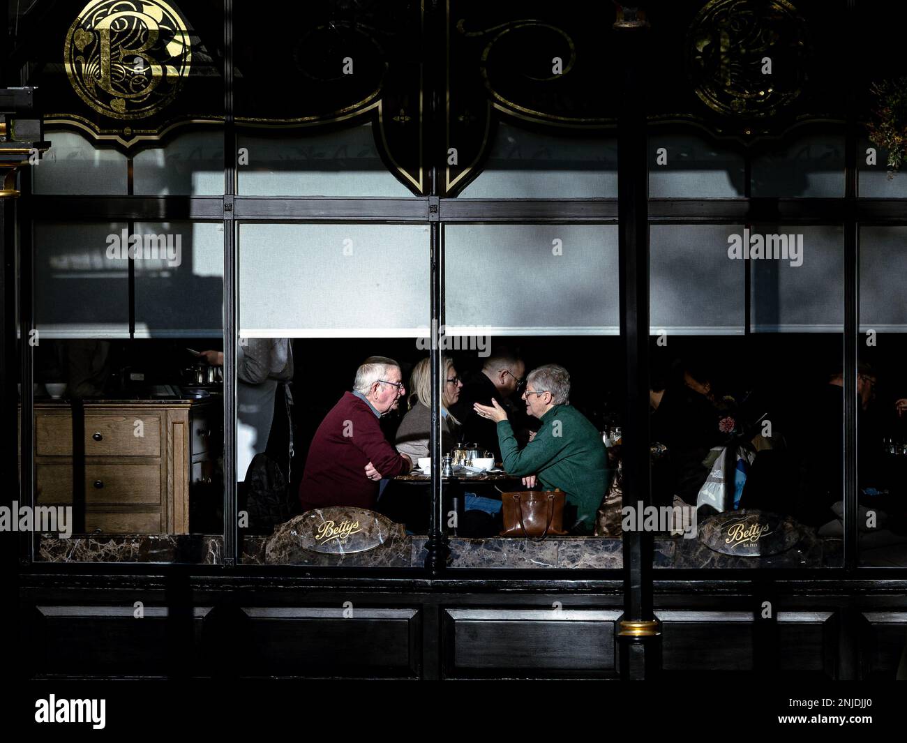 Un moment franc à l'intérieur du Bettys Café où deux personnes apprécient le thé et la conversation, capturés par la fenêtre avec une lumière naturelle douce illuminant le confortable Banque D'Images