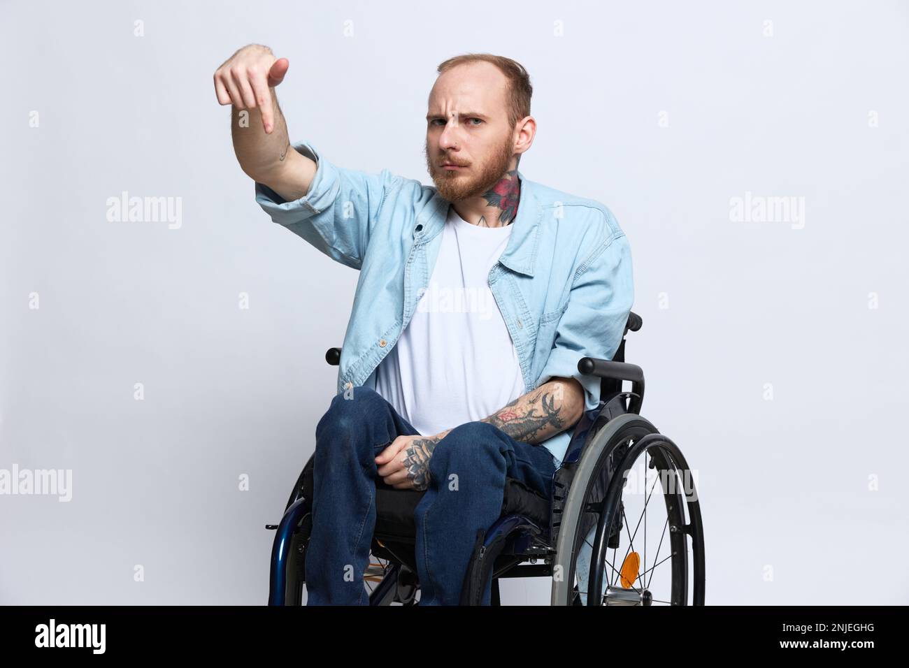 Un homme dans un fauteuil roulant et pointant un doigt, un espace de copie, avec des tatouages sur ses mains est assis sur un fond gris studio, concept de santé une personne avec Banque D'Images