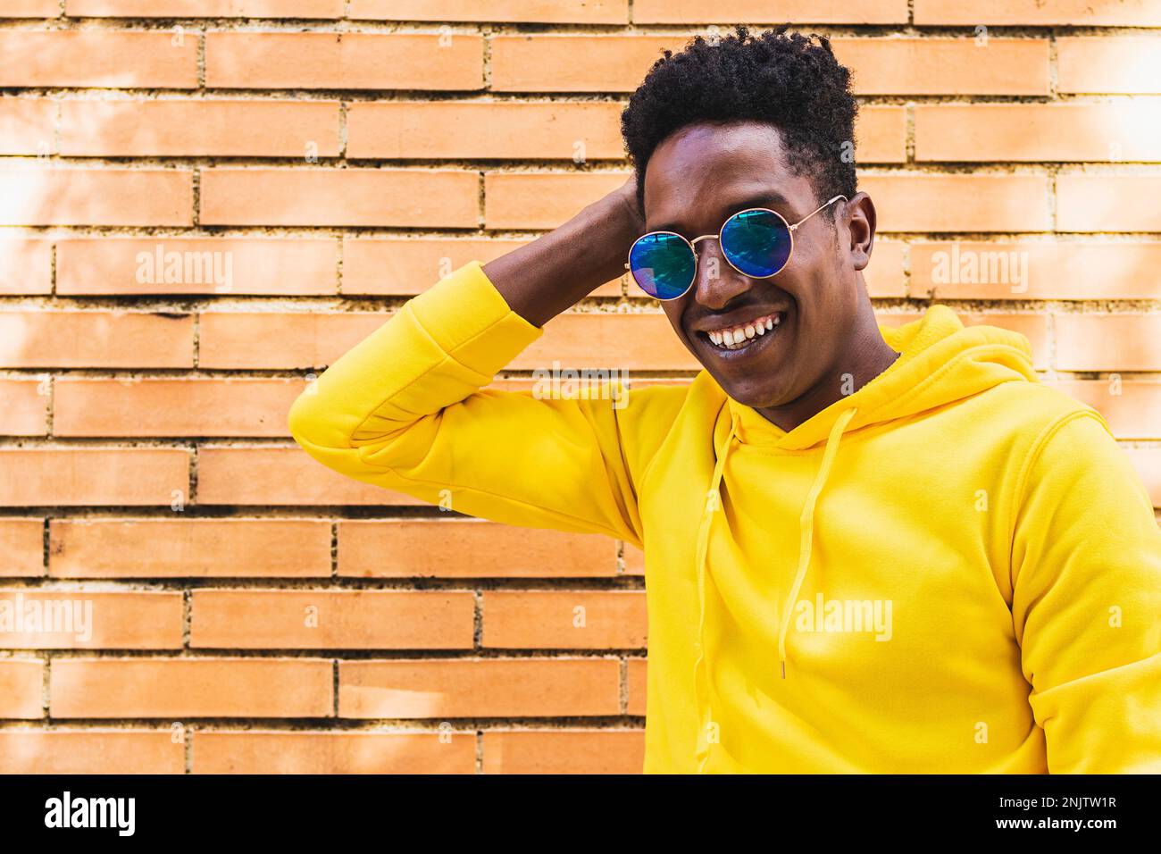 Homme africain souriant en pull à capuche jaune et lunettes de soleil debout contre le mur de brique rouge tout en touchant la tête Banque D'Images