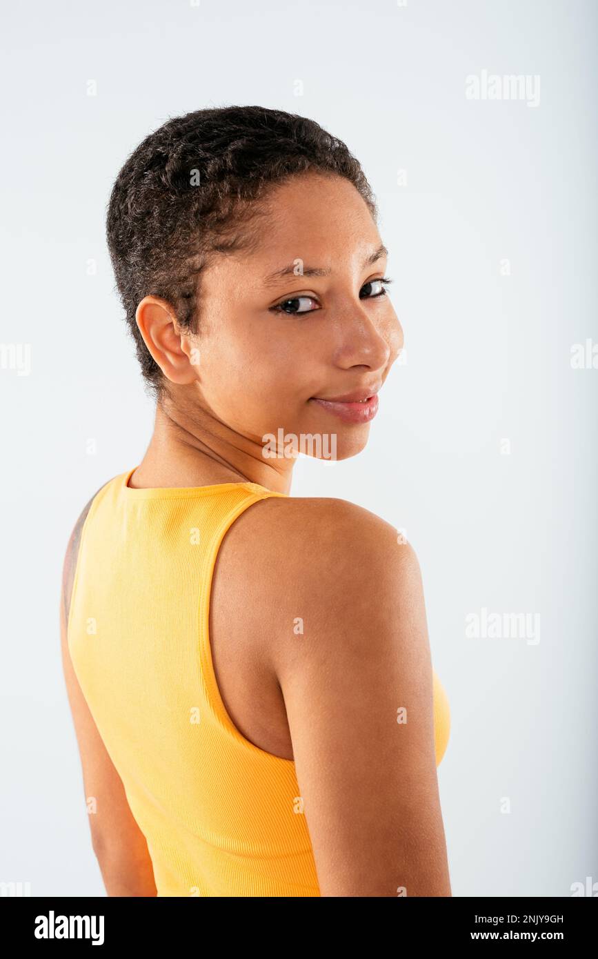 Vue arrière de positive joyeuse jeune afro-américaine avec des cheveux courts en jaune dessus et des jeans debout sur fond blanc regardant l'appareil photo Banque D'Images
