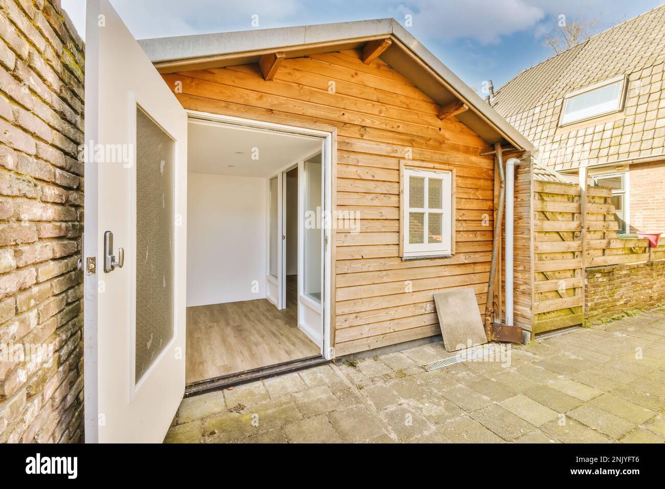 Extérieur de cour de petite maison rustique avec porte blanche ouverte et fenêtre avec façade en bois et carrelage en béton près du mur de briques Banque D'Images