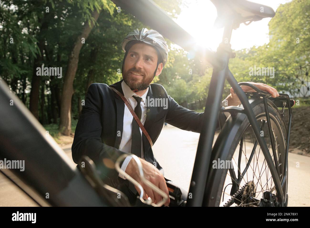 Homme souriant dans un costume élégant qui se déchort pour vérifier la roue de vélo dans le parc de la ville. Portrait d'un homme heureux entrepreneur en casque examinant la bicyclette, blanc Banque D'Images