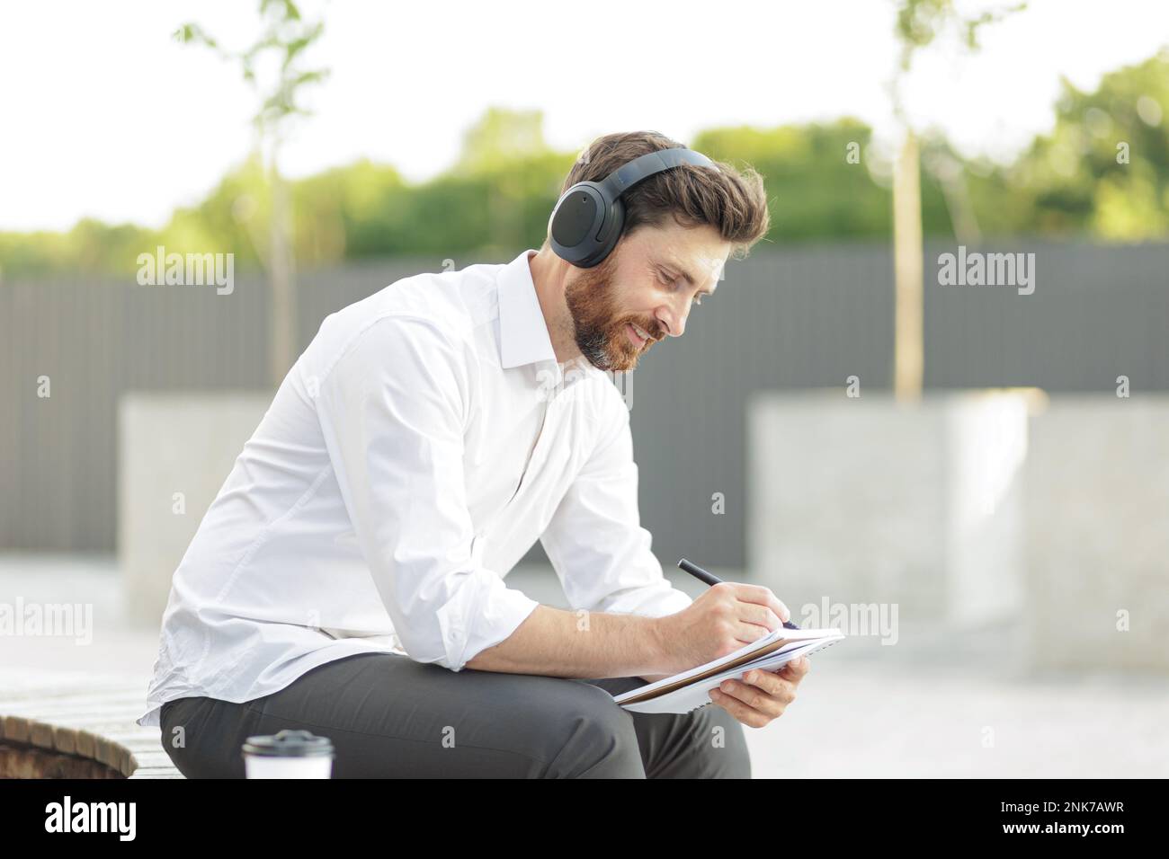 Vue latérale d'un employé de bureau caucasien dans un casque assis à l'extérieur tout en écrivant des idées créatives dans un ordinateur portable. Homme aux cheveux sombres qui écoute de la musique et du travail Banque D'Images