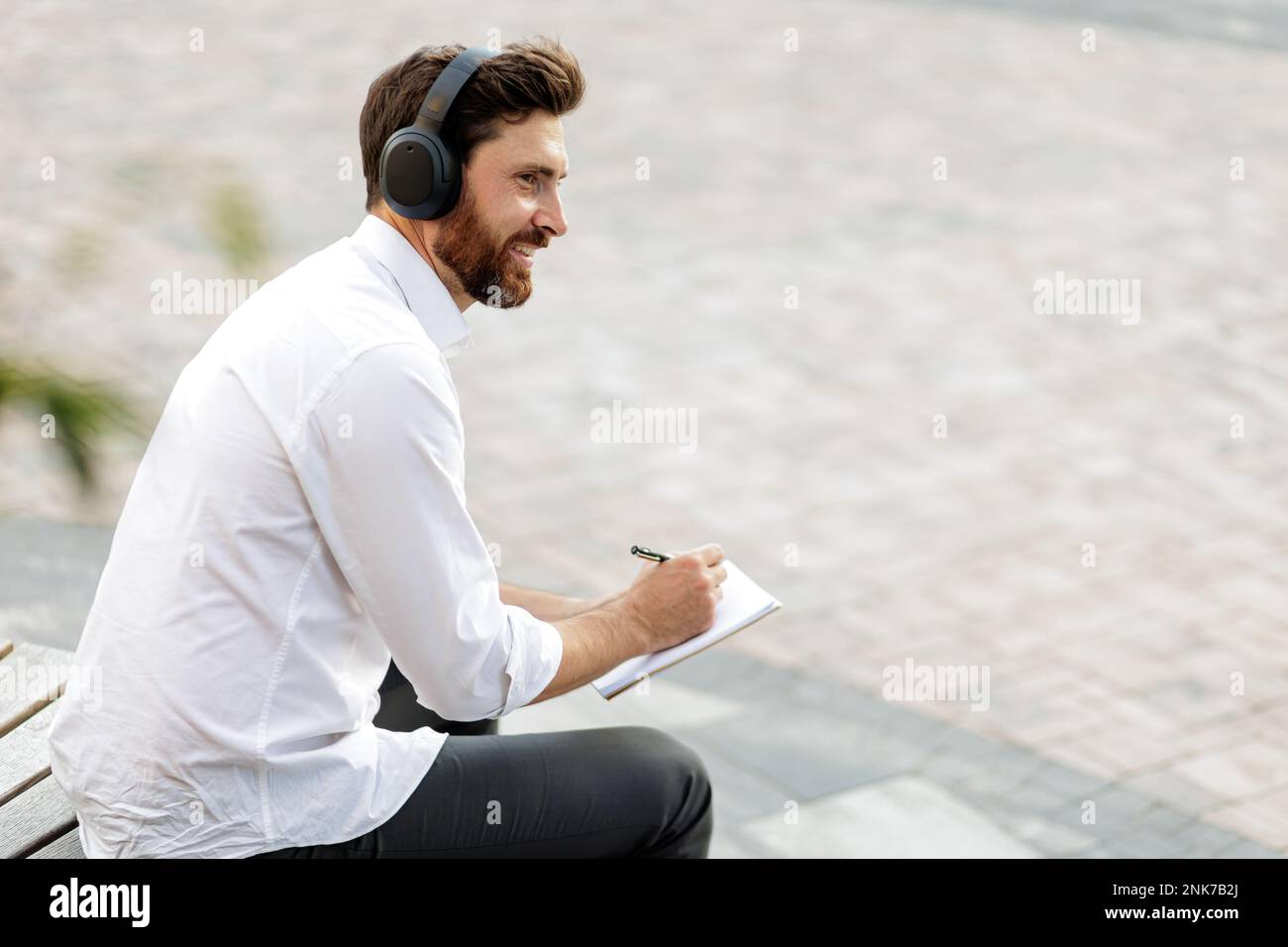 Homme d'affaires heureux écrivant de nouvelles idées dans un journal tout en écoutant de la musique dans un casque. Pigiste souriant avec piquant assis à l'extérieur dans un endroit calme et Banque D'Images