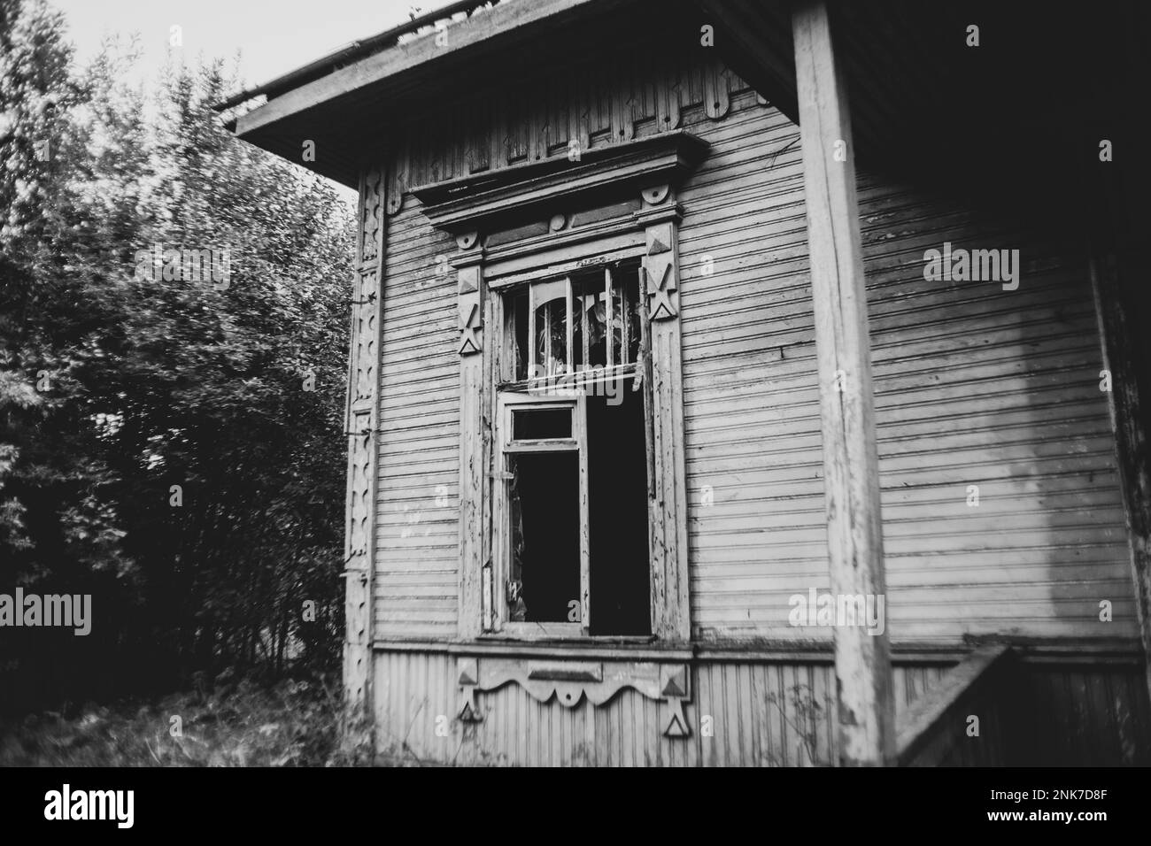 Une image en noir et blanc d'une maison de village en bois abandonnée avec des fenêtres cassées près des arbres. La vieille maison damée. Banque D'Images