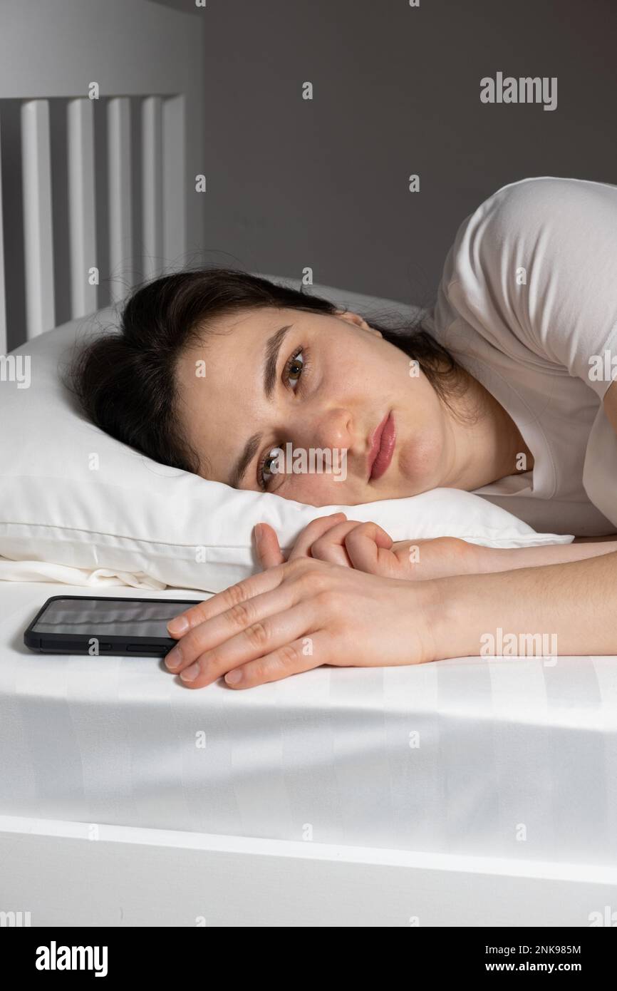 Une femme de 30 ans se trouve dans un lit avec un smartphone près d'un oreiller, addiction téléphonique Banque D'Images