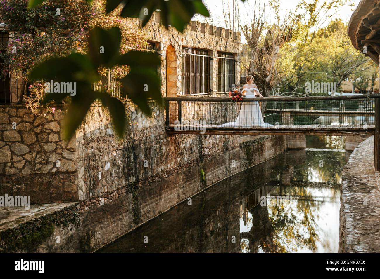 Belle mariée debout sur un pont rustique en bois au-dessus d'une rivière au coucher du soleil Banque D'Images