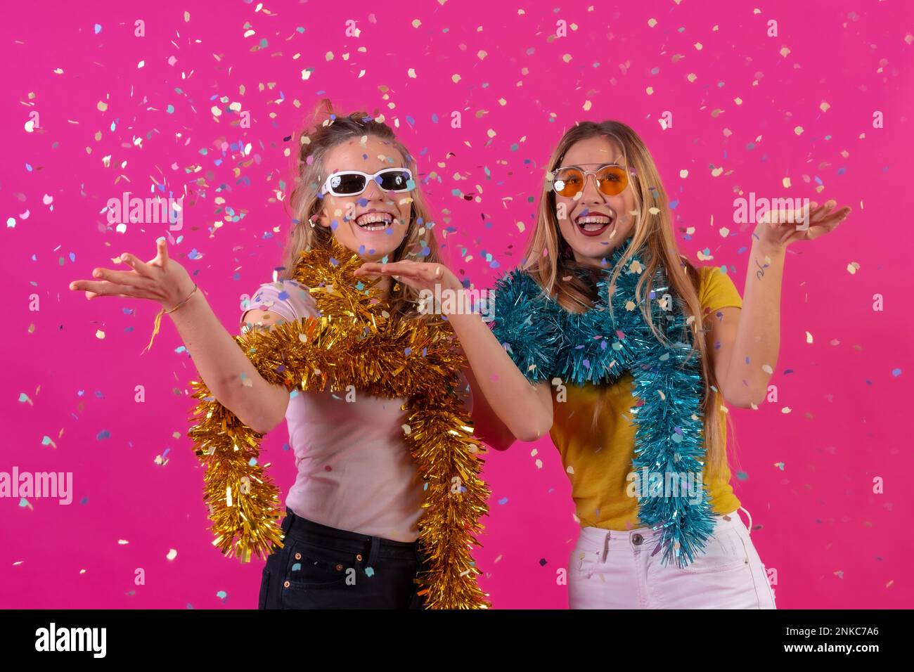 Deux jeunes femmes blanches blondes font la fête, à la discothèque, avec des confettis amusants à jeter, isolés sur fond rose Banque D'Images