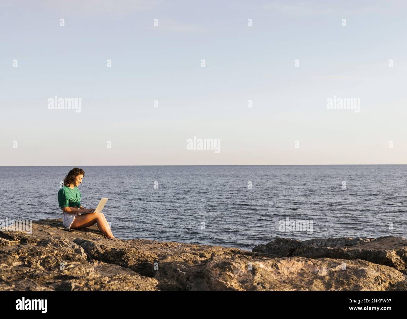 Femme travaillant sur un ordinateur portable assis sur le rocher au bord de la mer Banque D'Images