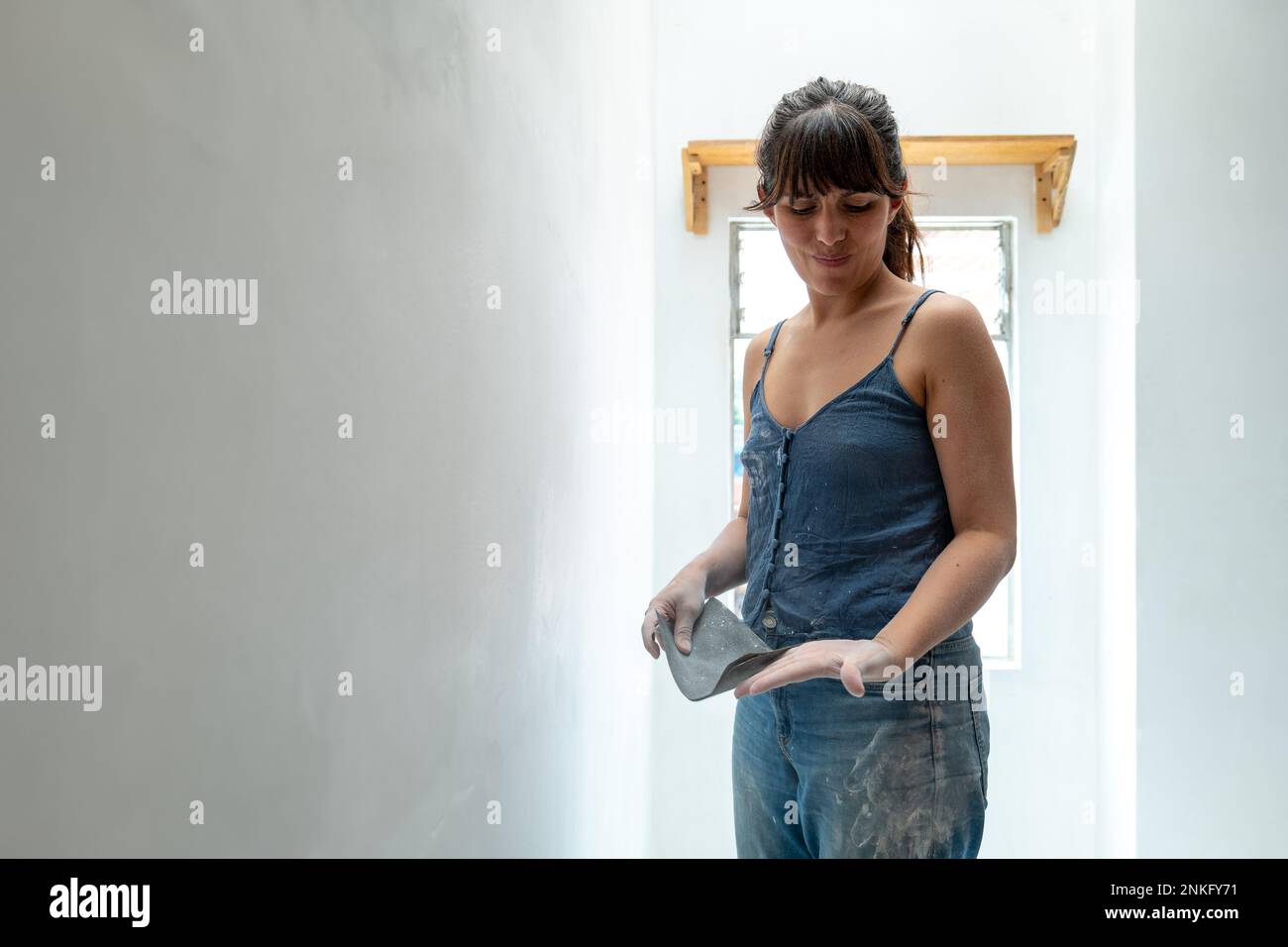 Une femme regarde ses mains sales en train de poncer un mur blanc avec du papier de verre Banque D'Images
