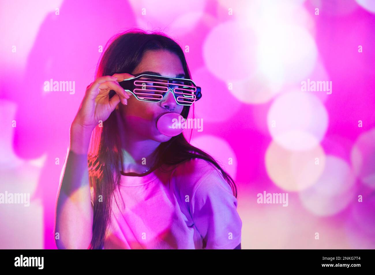 Femme portant des lunettes de fantaisie soufflant de la gomme de bulle sur fond de couleur néon Banque D'Images