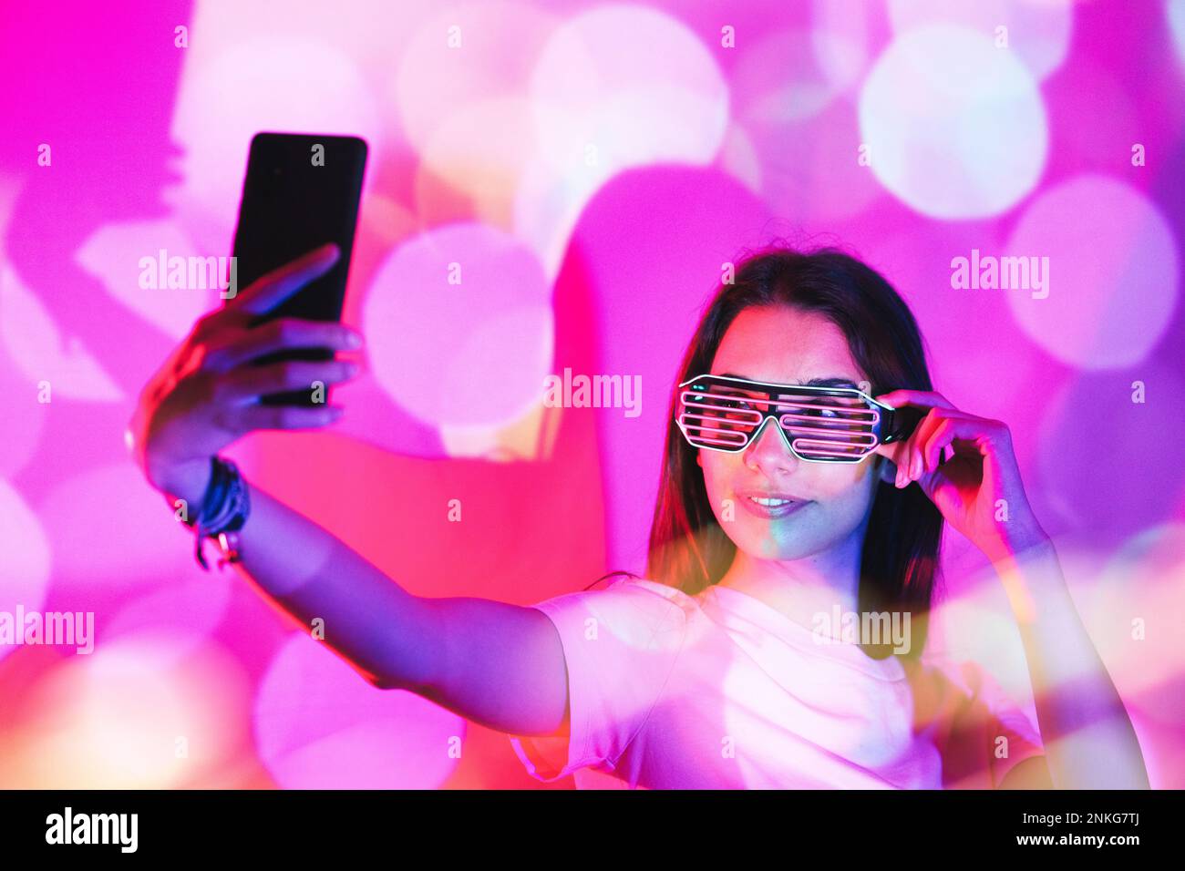 Jeune femme avec des lunettes de fantaisie prenant selfie sur smartphone dans des lumières néon roses Banque D'Images