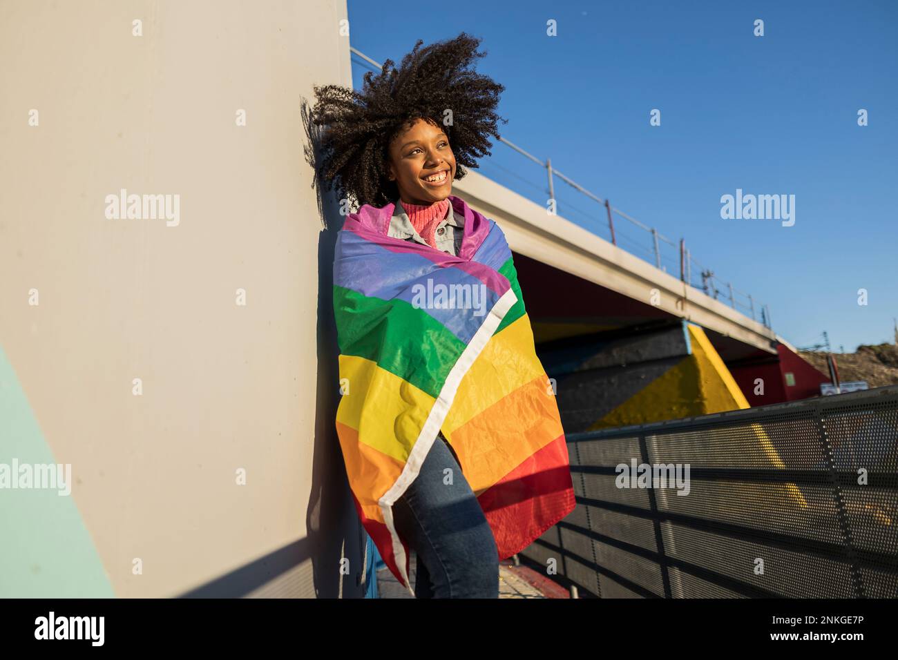 Jeune femme souriante enveloppée d'un drapeau arc-en-ciel appuyé sur le mur Banque D'Images