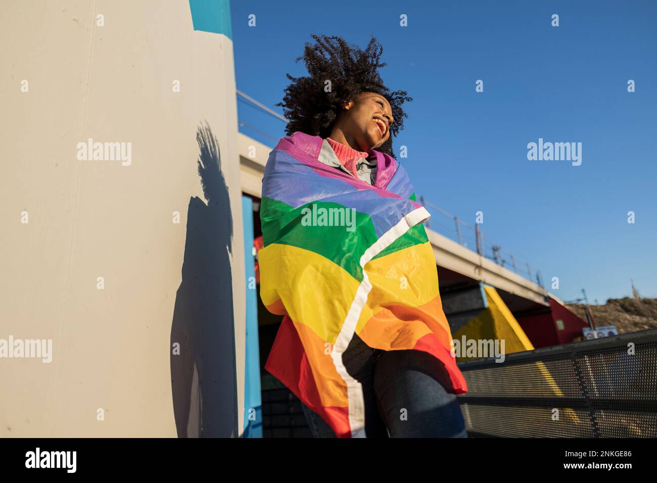 Jeune femme joyeuse enveloppée de drapeau arc-en-ciel en appréciant le soleil Banque D'Images