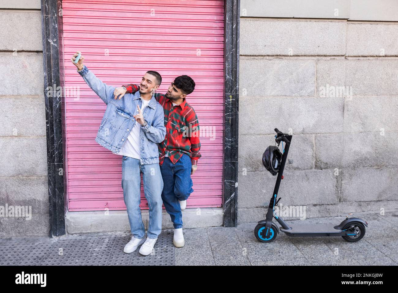 Un jeune couple gay prend un selfie à l'aide d'un smartphone devant le mur du volet roulant Banque D'Images