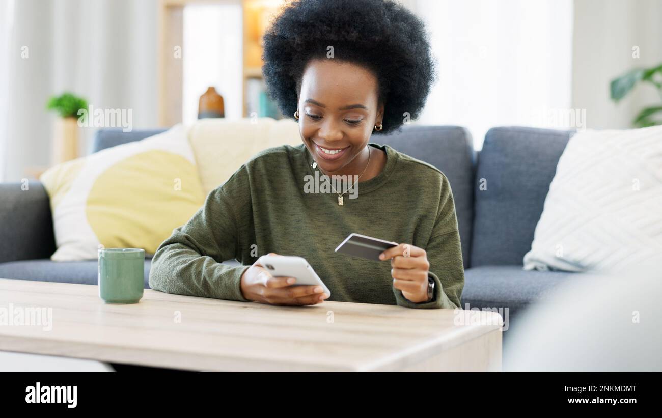 Jeune femme noire magasiner en ligne à l'aide de son téléphone et de sa carte bancaire dans un salon lumineux. Services bancaires et gestion des actifs et des finances à l'aide de Banque D'Images