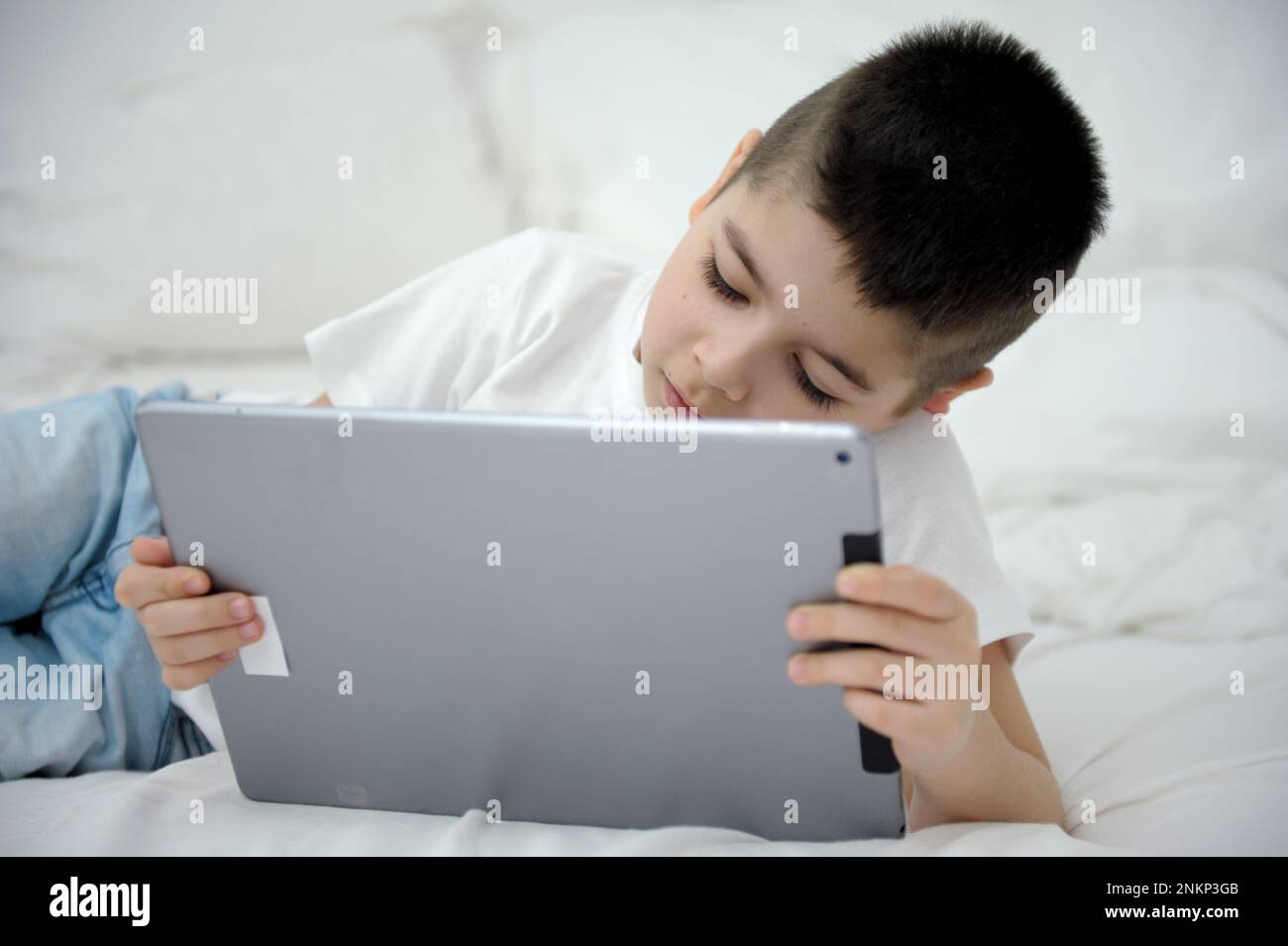 Un petit garçon de 7 ans se couche sur le lit dans les mains avec la tablette en ligne d'apprentissage intéressant-film applications de jeux mobiles Je suis seul à la maison passer du temps sur les réseaux sociaux Internet les enfants et l'ordinateur Banque D'Images