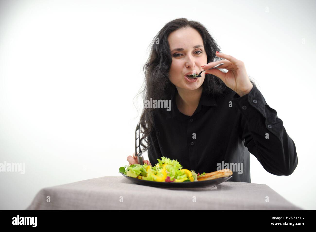 sur fond blanc, une femme en vêtements noirs mange une salade et de la viande avec une fourchette regarde dans l'espace cadre pour le texte bonne nutrition fond blanc une femme sans goût alimentation forcée Banque D'Images