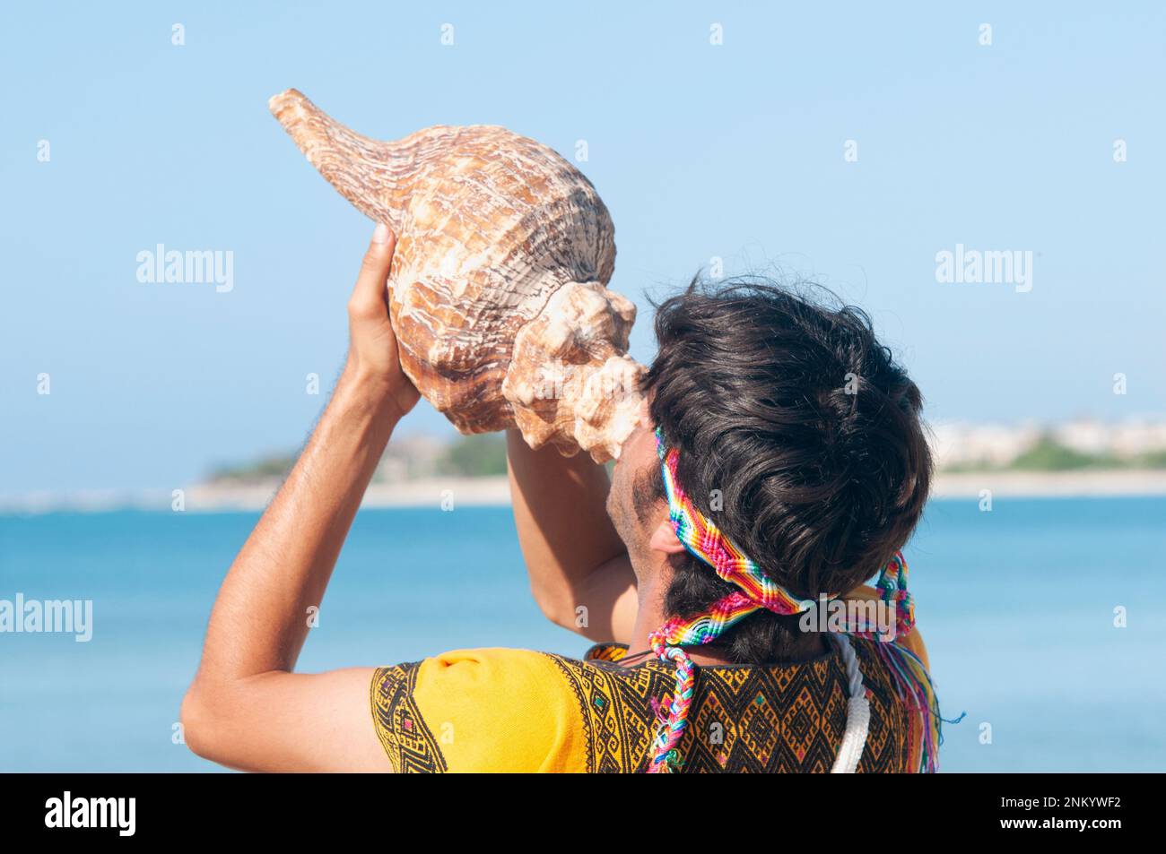 Homme maya en costume traditionnel jouant dans une carapace de mer sur la plage pendant une cérémonie. Vue arrière Banque D'Images
