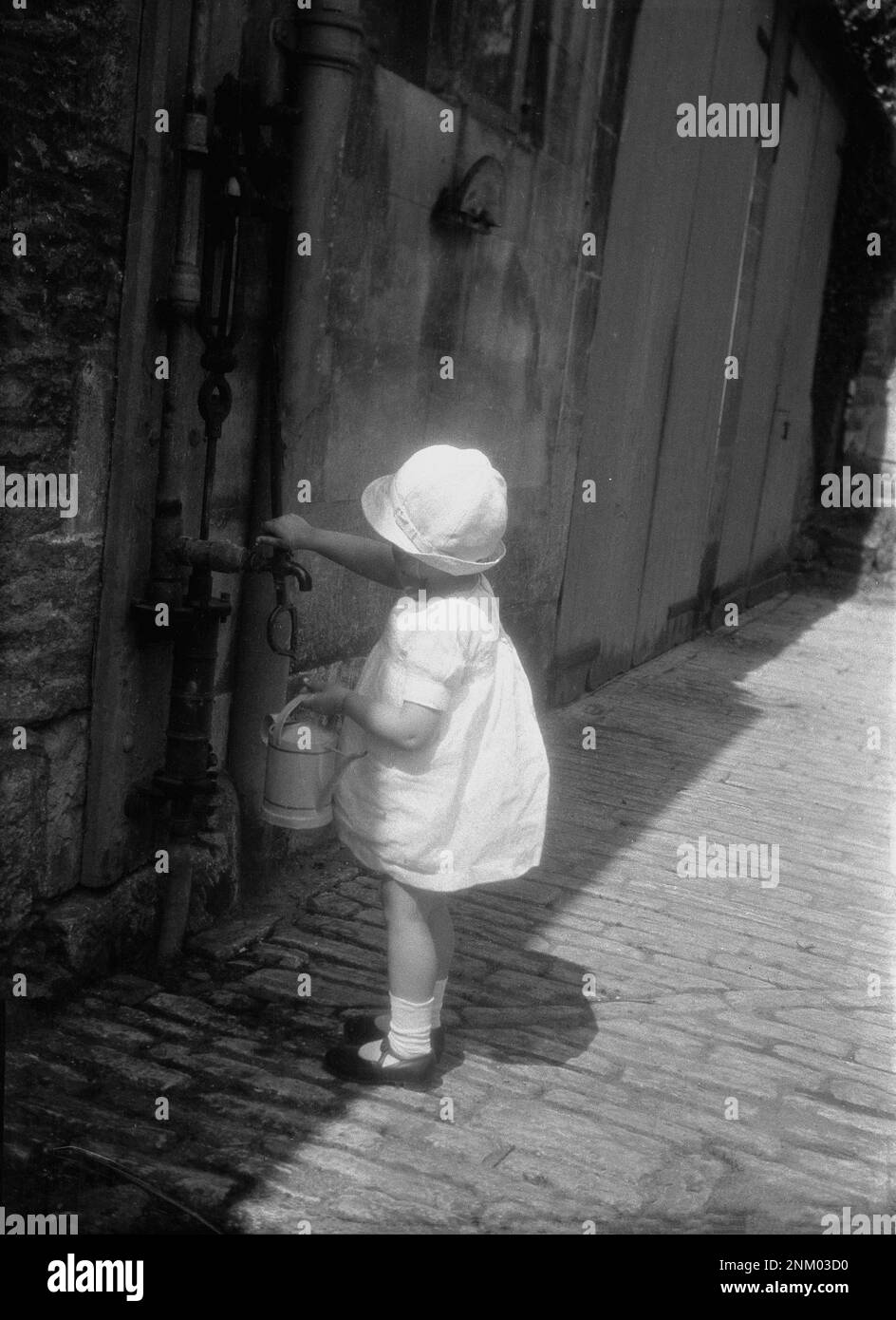 1931, historique, l'été et à l'extérieur d'une petite fille dans une robe en coton et un bonnet de soleil, sur le point de remplir son jouet arrosoir, à un robinet d'eau extérieur, Angleterre, Royaume-Uni. Banque D'Images